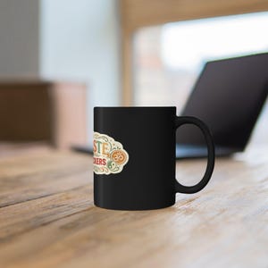 The black "Namaste Motherfuckers" funny yoga coffee mug by SarcasmArt on a wooden desk in a bright home office.