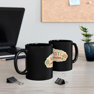 Two black "Namaste Motherfuckers" funny yoga coffee mug by SarcasmArts on an office desk next to a laptop – a sarcastic gift for yoga lovers and coworkers.