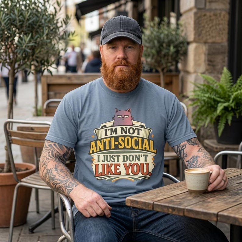 Bearded man in a cap with a coffee at a café table wearing the "I'm Not Anti-Social, I Just Don't Like You" sarcastic introvert T-shirt by SarcasmArt in Blue Jean.