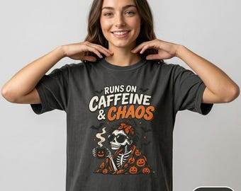 Op de afbeelding: Een antracietgrijs t-shirt met een afbeelding van een skelet met een zonnebril en een deken met pompoenpatroon, dat een dampende mok vasthoudt. De tekst "RUNS ON CAFFEINE & CHAOS" staat in witte en oranje letters, omringd door kleine vleermuizen.