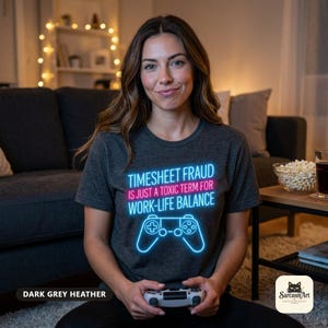 Woman holding a game controller in a cozy living room wearing the "Timesheet Fraud Is Just a Toxic Term for Work-Life Balance" neon gamer T-shirt by SarcasmArt in Dark Grey Heather.