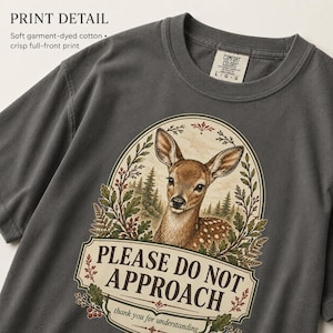 May include: A charcoal gray short-sleeve t-shirt featuring a graphic of a fawn inside an oval frame with forest elements. The text reads PLEASE DO NOT APPROACH and thank you for understanding. The shirt has a crew neck and a size L label.