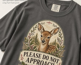May include: A charcoal gray short-sleeve t-shirt featuring a graphic of a fawn inside an oval frame with forest elements. The text reads PLEASE DO NOT APPROACH and thank you for understanding. The shirt has a crew neck and a size L label.