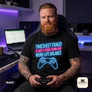 Bearded gamer with a controller at his gaming setup wearing the "Timesheet Fraud Is Just a Toxic Term for Work-Life Balance" neon gamer T-shirt by SarcasmArt in Black – a Bella+Canvas 3001 unisex tee with glowing neon blue and pink lettering TIMESHEET FRAUD IS JUST A TOXIC TERM FOR WORK-LIFE BALANCE above a neon game controller.