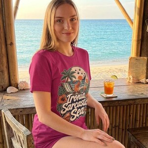 Woman sitting at a tropical beach bar wearing the raspberry pink "Ironia, Sarcastic Sea" retro beach T-shirt by SarcasmArt, a funny summer vacation shirt.