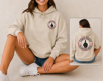 May include: A beige hooded sweatshirt featuring a circular graphic on the chest and back. The graphic displays a hand making a middle finger gesture surrounded by the text "LIEBER ASOZIAL ALS GAR NICHT SOZIAL" in a red and black circular border.