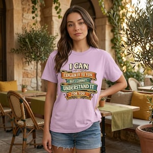 May include: A light purple short-sleeve t-shirt featuring a graphic with the text I CAN EXPLAIN IT TO YOU, BUT I CANNOT UNDERSTAND IT FOR YOU. The design includes layered orange, teal, and yellow banners with decorative flourishes.