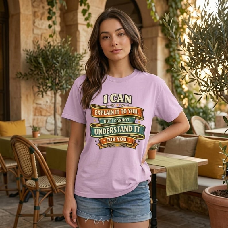 Woman in a sunny café courtyard wearing the "I Can Explain It to You, But I Can't Understand It for You" sarcastic teacher T-shirt by SarcasmArt in Orchid (soft pink) – a Comfort Colors 1717 unisex tee with vintage ribbon banners reading I CAN EXPLAIN IT TO YOU BUT I CAN'T UNDERSTAND IT FOR YOU in orange, green and cream.