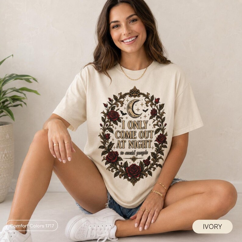 Smiling woman sitting on the floor wearing the "I Only Come Out at Night" romantic gothic vampire T-shirt by SarcasmArt in Ivory.