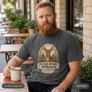 Bearded man with a coffee cup on a sunny porch wearing the "Please Do Not Approach" vintage fawn introvert T-shirt by SarcasmArt in Pepper (washed charcoal).