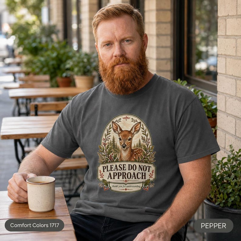 Bearded man with a coffee cup on a sunny porch wearing the "Please Do Not Approach" vintage fawn introvert T-shirt by SarcasmArt in Pepper (washed charcoal).