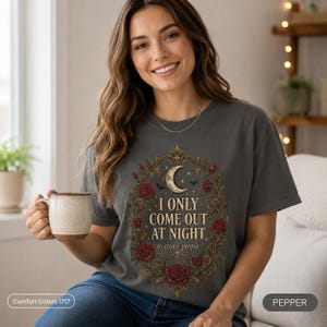 Woman holding a coffee mug wearing the "I Only Come Out at Night" romantic gothic vampire T-shirt by SarcasmArt in Pepper (washed charcoal) – a dark academia and gothic gift.