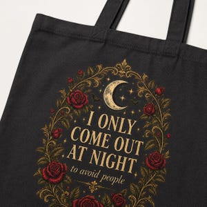 Print detail of the Black "I Only Come Out at Night to Avoid People" gothic canvas tote bag by SarcasmArt: a crescent moon and dark red Victorian roses in a gold filigree frame around the words I ONLY COME OUT AT NIGHT to avoid people.