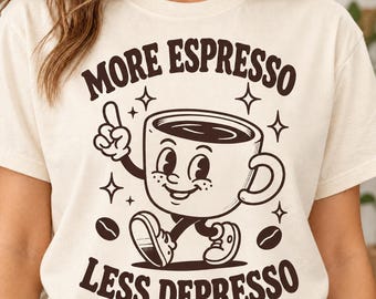 May include: A cream-colored t-shirt featuring a dark brown graphic of a smiling coffee cup character with arms and legs. The text above reads MORE ESPRESSO and the text below reads LESS DEPRESSO, surrounded by small star and coffee bean icons.