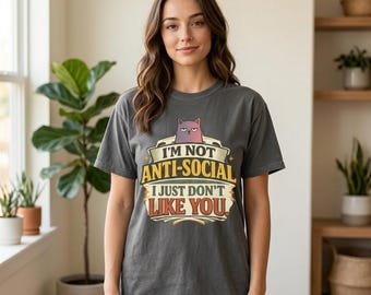 Op de afbeelding: Een antracietgrijs t-shirt met korte mouwen en een afbeelding van een norse paarse kat boven de tekst "I'M NOT ANTI-SOCIAL I JUST DON'T LIKE YOU." Het ontwerp bevat gele en witte letters in een vintage-geïnspireerde lay-out.