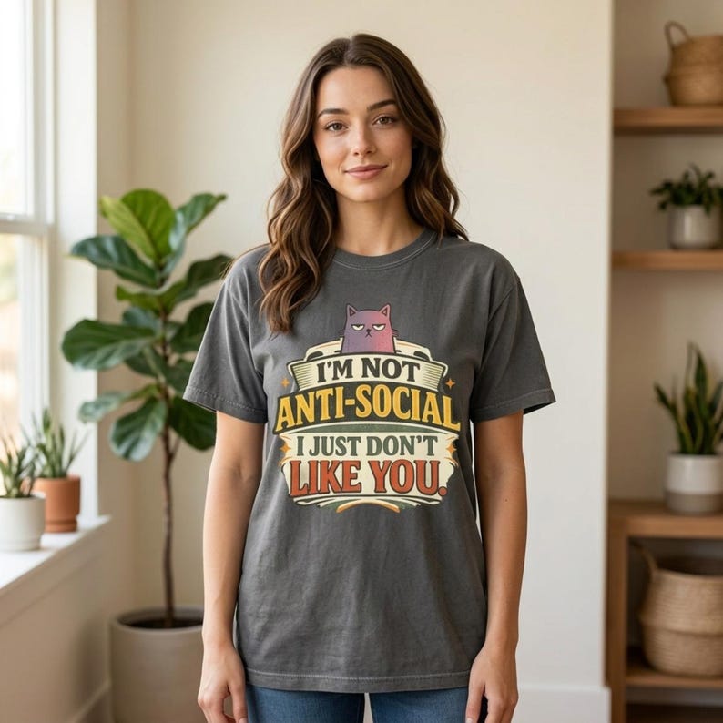 Woman in a bright room with plants wearing the "I'm Not Anti-Social, I Just Don't Like You" sarcastic introvert T-shirt by SarcasmArt in Pepper (washed charcoal) – a Comfort Colors 1717 unisex tee with a grumpy cat peeking over bold retro banner lettering reading I'M NOT ANTI-SOCIAL I JUST DON'T LIKE YOU in yellow, cream and red.