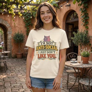Smiling woman in a sunny courtyard café wearing the "I'm Not Anti-Social, I Just Don't Like You" sarcastic introvert T-shirt by SarcasmArt in Ivory.
