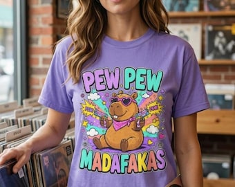 May include: A light purple t-shirt featuring a graphic of a brown capybara wearing heart-shaped sunglasses and a pink bandana, holding two toy guns. The text reads PEW PEW MADAFAKAS in colorful letters, surrounded by clouds, stars, and hearts.