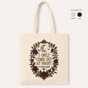 May include: A natural-coloured canvas tote bag featuring a central graphic of a crescent moon, small bats, and stars surrounded by a detailed ornate frame of red roses and green leaves. The text reads, I ONLY COME OUT AT NIGHT to avoid people.