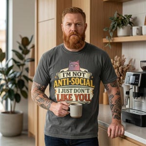 Bearded man holding a mug in a kitchen wearing the "I'm Not Anti-Social, I Just Don't Like You" sarcastic introvert T-shirt by SarcasmArt in Pepper.