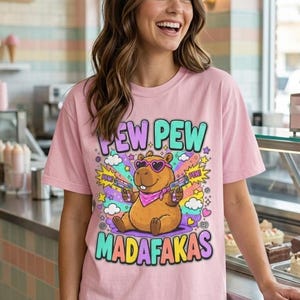 Smiling woman in a café wearing the "Pew Pew Madafakas" capybara meme T-shirt by SarcasmArt in Blossom (soft pink).