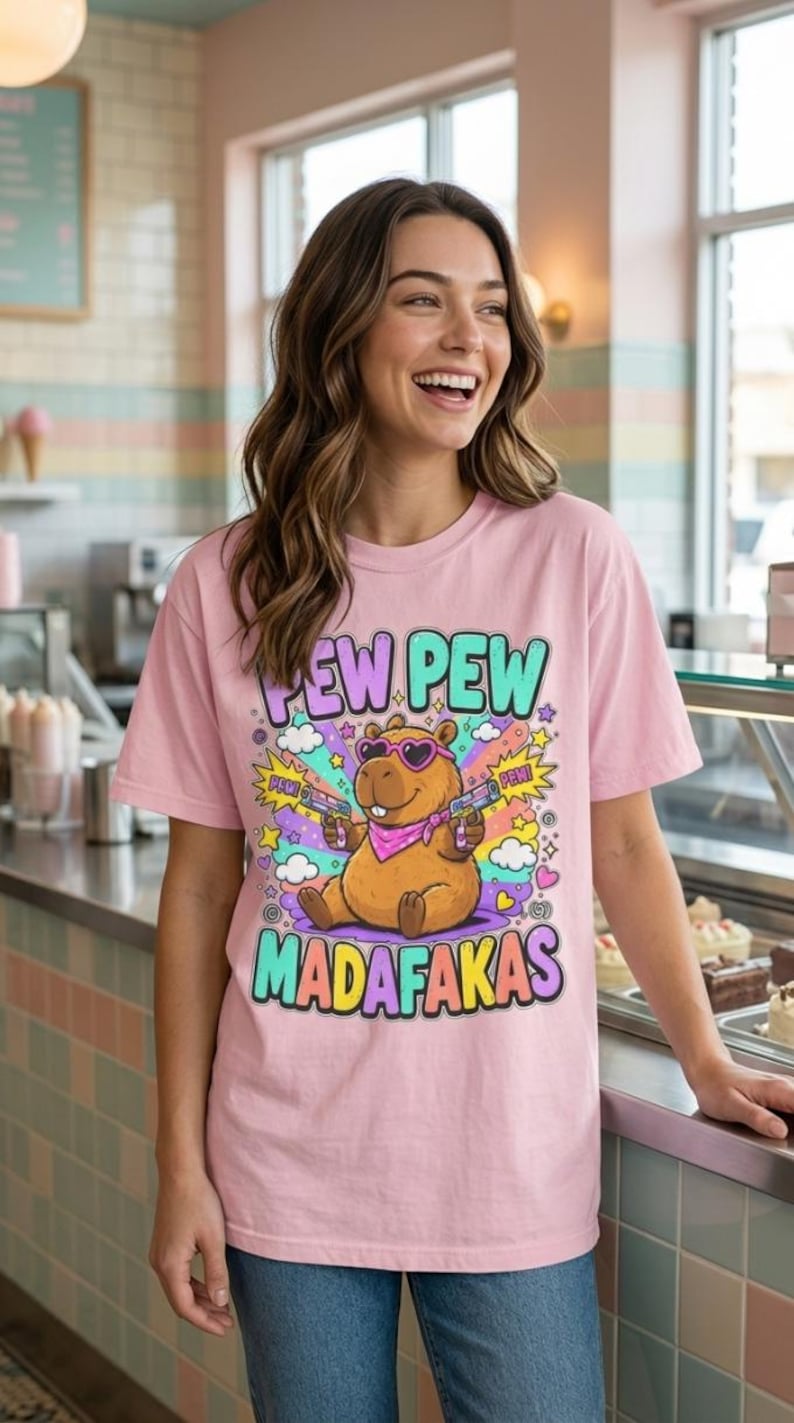 Smiling woman in a café wearing the "Pew Pew Madafakas" capybara meme T-shirt by SarcasmArt in Blossom (soft pink).