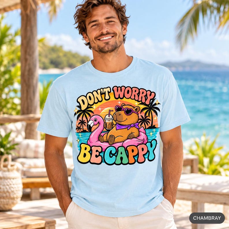 Smiling man on a tropical beach wearing the "Don't Worry Be Cappy" capybara beach T-shirt by SarcasmArt in Chambray (light blue).