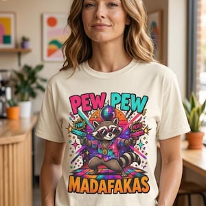 Woman in a sunny loft wearing the "Pew Pew Madafakas" raccoon meme T-shirt by SarcasmArt in Ivory – a Comfort Colors 1717 unisex tee with a neon trash panda raccoon with laser guns, paint splashes and bold PEW PEW MADAFAKAS lettering in pink, teal and orange.