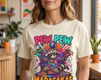 May include: A cream-colored t-shirt featuring a graphic of a raccoon wearing sunglasses and a patterned shirt, holding two toy guns. Above the raccoon is a disco ball and the text PEW PEW, with the text MADAFAKAS printed in orange below.