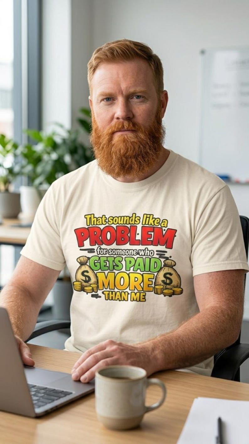 Bearded man at his desk with a laptop and mug wearing the "That Sounds Like a Problem for Someone Who Gets Paid More Than Me" sarcastic coworker T-shirt by SarcasmArt in Ivory.