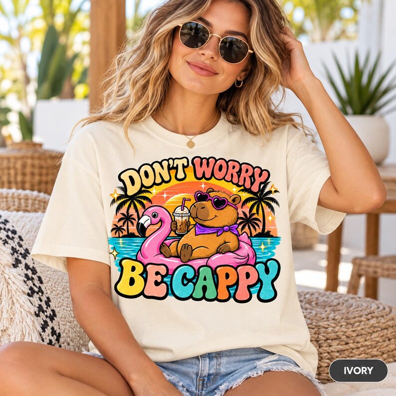 Woman in sunglasses sitting on a rattan chair wearing the "Don't Worry Be Cappy" capybara beach T-shirt by SarcasmArt in Ivory.