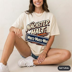 May include: A natural-coloured short-sleeve t-shirt featuring a graphic design with the text NÄCHSTER HALT and a blue rectangular sign graphic containing the text Merz-Muss-Weg in white lettering with a distressed texture and orange accents.