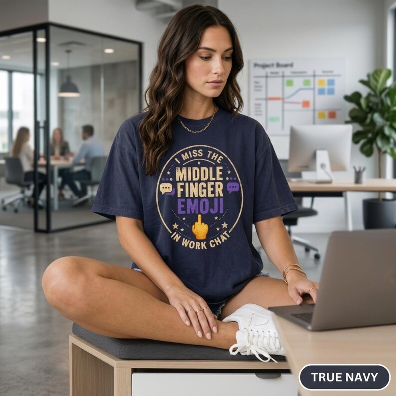 Woman sitting on a desk in a modern office wearing the "I Miss the Middle Finger Emoji in Work Chat" sarcastic office T-shirt by SarcasmArt in True Navy – a funny remote work and coworker gift.