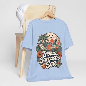 The "Ironia, Sarcastic Sea" retro beach T-shirt by SarcasmArt in light blue hanging on a wooden chair.