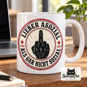 May include: A white ceramic mug featuring a circular graphic with a distressed red border and two red stars. The design displays a black silhouette of a hand making a middle finger gesture, surrounded by the text LIEBER ASOZIAL ALS GAR NICHT SOZIAL.