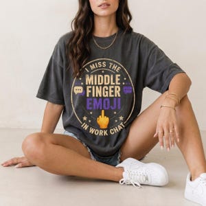 Könnte beinhalten: Ein anthrazitfarbenes Kurzarm-T-Shirt mit einem kreisförmigen Aufdruck mit dem Text I MISS THE MIDDLE FINGER EMOJI IN WORK CHAT. Das Design enthält ein gelbes Mittelfinger-Emoji, violette Sprechblasen und kleine Sterne im Used-Look.