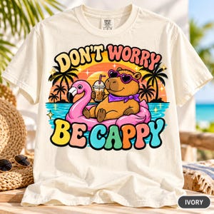 Ivory "Don't Worry Be Cappy" capybara beach T-shirt by SarcasmArt on a beach lounge chair – Comfort Colors 1717 unisex tee with a relaxed capybara in sunglasses floating on a pink flamingo ring with a cocktail, palm trees and a retro sunset, with colorful DON'T WORRY BE CAPPY lettering.