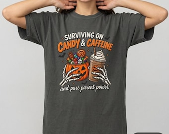 May include: A charcoal gray t-shirt featuring a graphic of two skeleton hands holding a jack-o'-lantern bucket filled with candy and a coffee cup. The text reads "SURVIVING ON CANDY & CAFFEINE" and "and pure parent power" in orange and white.