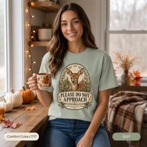 Woman holding a cup of tea in a cozy autumn kitchen wearing the "Please Do Not Approach" vintage fawn introvert T-shirt by SarcasmArt in Bay (sage green) – a funny homebody gift.