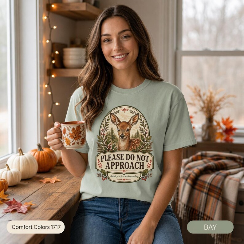 Woman holding a cup of tea in a cozy autumn kitchen wearing the "Please Do Not Approach" vintage fawn introvert T-shirt by SarcasmArt in Bay (sage green) – a funny homebody gift.