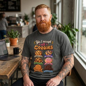 Bearded man in a kitchen wearing the "Yes, I Accept All Cookies" funny programmer T-shirt by SarcasmArt in Pepper (washed charcoal) – a funny tech gift for developers and IT engineers.