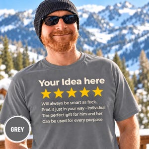 Bearded man in a beanie and sunglasses in front of snowy mountains wearing the personalized golden 5-star review T-shirt by SarcasmArt in Grey, with your custom title, five golden stars and four lines of your own review text in white.