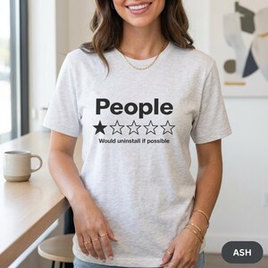Woman in a bright kitchen wearing the "People – 1 Star, Would Uninstall If Possible" funny review T-shirt by SarcasmArt in Ash (light heather grey).