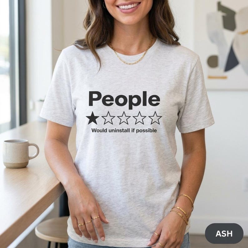 Woman in a bright kitchen wearing the "People – 1 Star, Would Uninstall If Possible" funny review T-shirt by SarcasmArt in Ash (light heather grey).