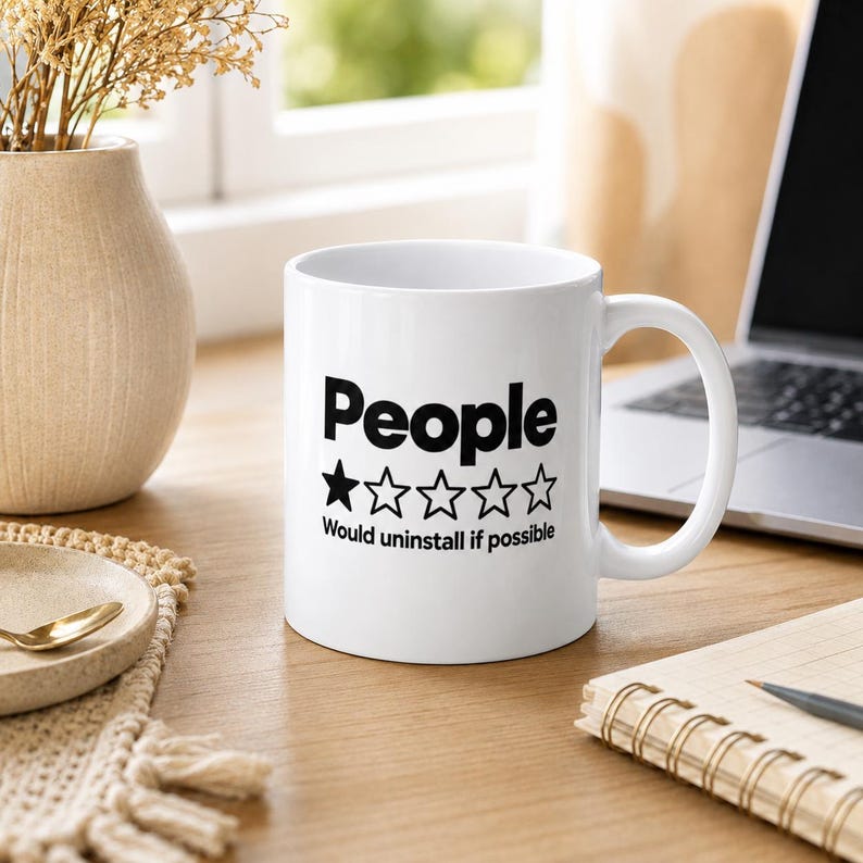 The "People – 1 Star, Would Uninstall If Possible" sarcastic coffee mug by SarcasmArt on a desk next to a laptop and notebook, a minimalist office humor mug.