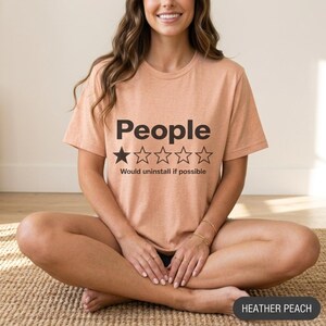 Woman sitting cross-legged wearing the "People – 1 Star, Would Uninstall If Possible" funny review T-shirt by SarcasmArt in Heather Peach.