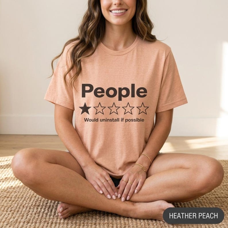 Woman sitting cross-legged wearing the "People – 1 Star, Would Uninstall If Possible" funny review T-shirt by SarcasmArt in Heather Peach.