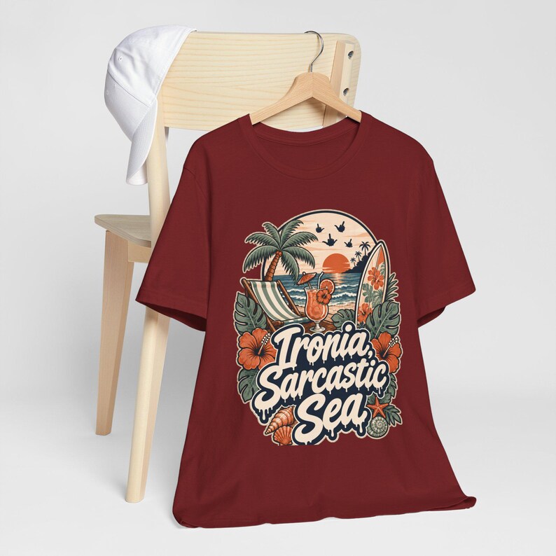 The "Ironia, Sarcastic Sea" retro beach T-shirt by SarcasmArt in maroon hanging on a wooden chair.