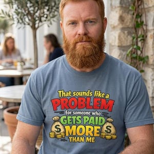 Bearded man at a café table wearing the "That Sounds Like a Problem for Someone Who Gets Paid More Than Me" sarcastic coworker T-shirt by SarcasmArt in Blue Jean.