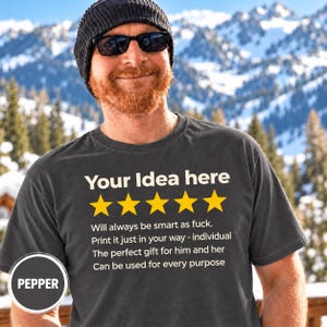 Bearded man in a beanie and sunglasses in front of snowy mountains wearing the personalized golden 5-star review T-shirt by SarcasmArt in Pepper (washed charcoal), with your custom title, five golden stars and four lines of your own review text in white.